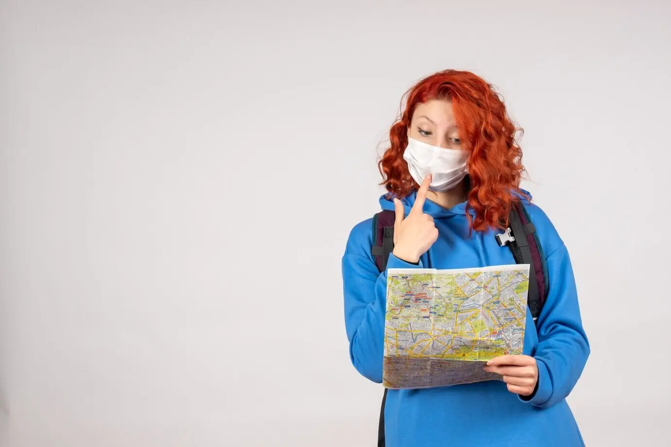 Vorderansicht einer jungen Frau mit Rucksack und Karte, die eine Maske trägt, vor einer weißen Wand.